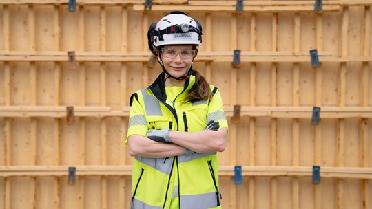 Työnjohtoharjoittelija työmaalla henkilösuojaimet päällä puisen seinän edessä.